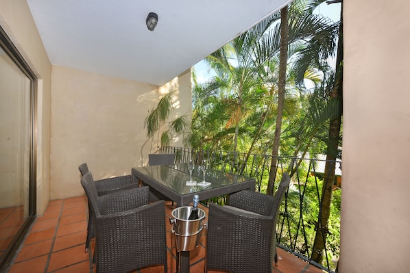 A balcony with a glass table and wicker chairs, overlooking a tropical garden.