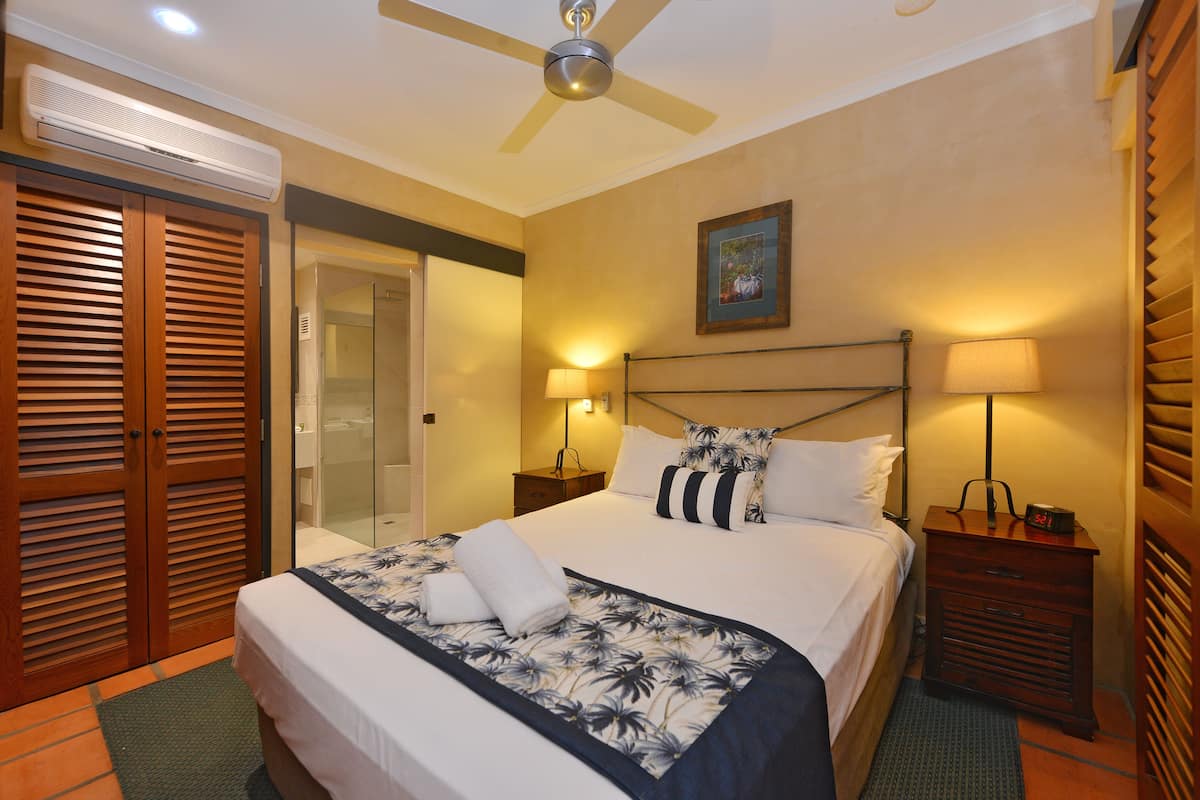 Une chambre d’hôtel comprenant un lit, une table de chevet, une lampe, un ventilateur de plafond et une porte avec des volets à persiennes.