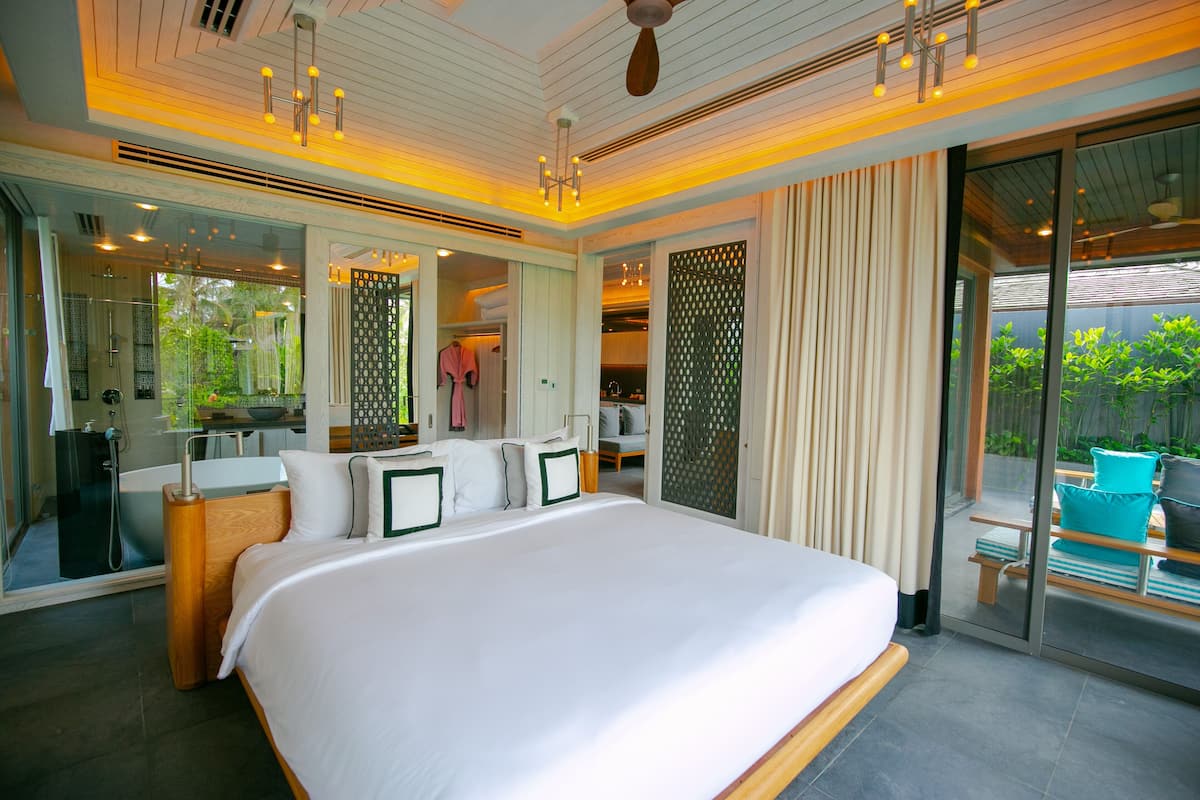 A modern hotel room with a large bed, a bathroom with a bathtub, and a view of a garden through large windows.
