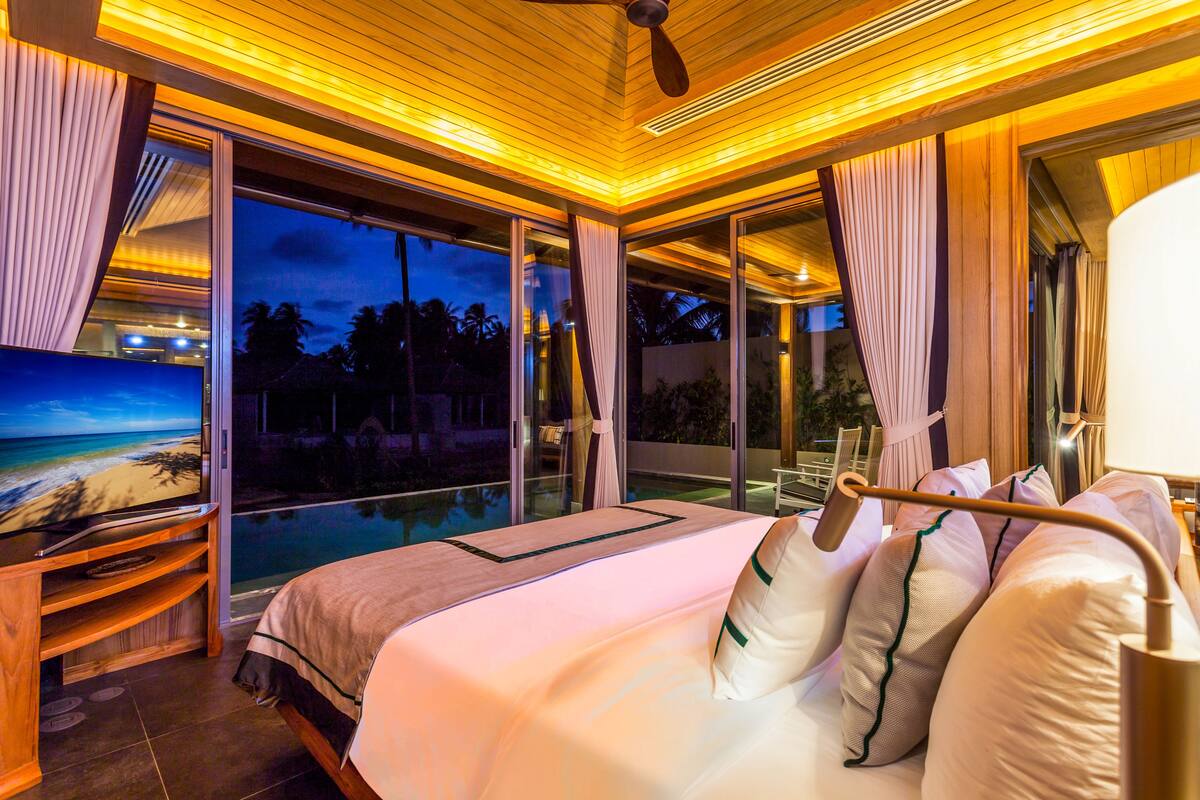 A bedroom with a large bed, a television, and a view of a pool and palm trees.