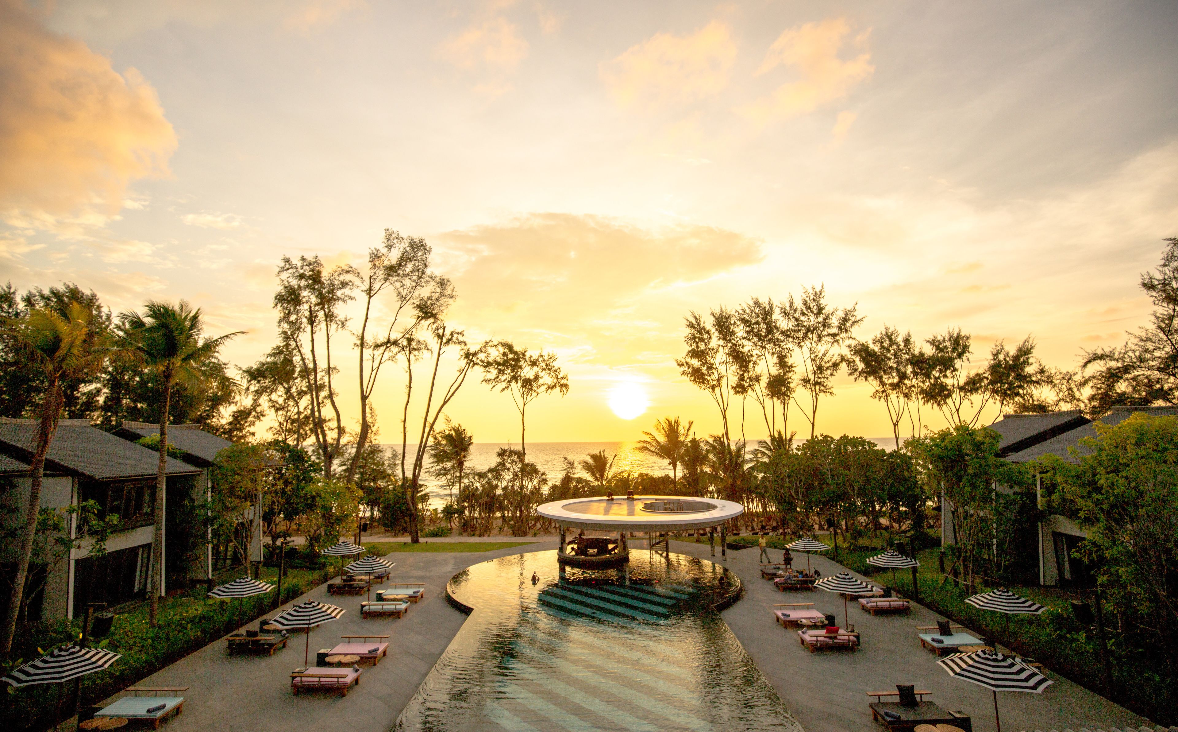 A resort pool area with lounge chairs and umbrellas, surrounded by palm trees and a sunset view.