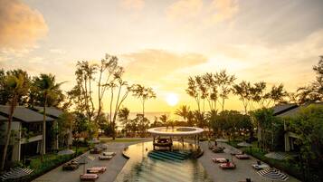 A resort pool area with lounge chairs and umbrellas, surrounded by palm trees and a sunset view.