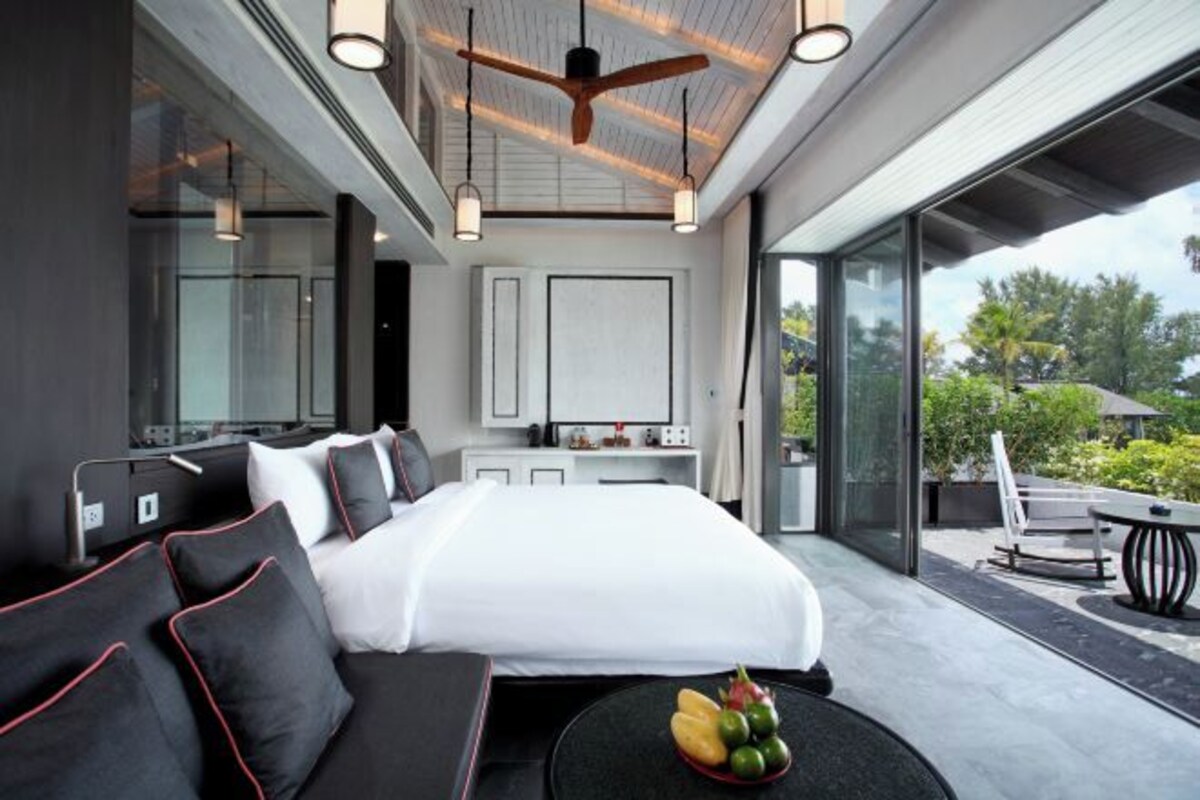 Ein modernes Hotelzimmer mit einem großen Bett, einer Couch, einem Esstisch mit Obst und Blick ins Freie.