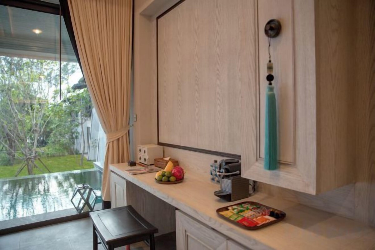 Une cuisine donnant sur un jardin grâce à une fenêtre, un plan de travail avec un panier de fruits, et un objet décoratif suspendu.