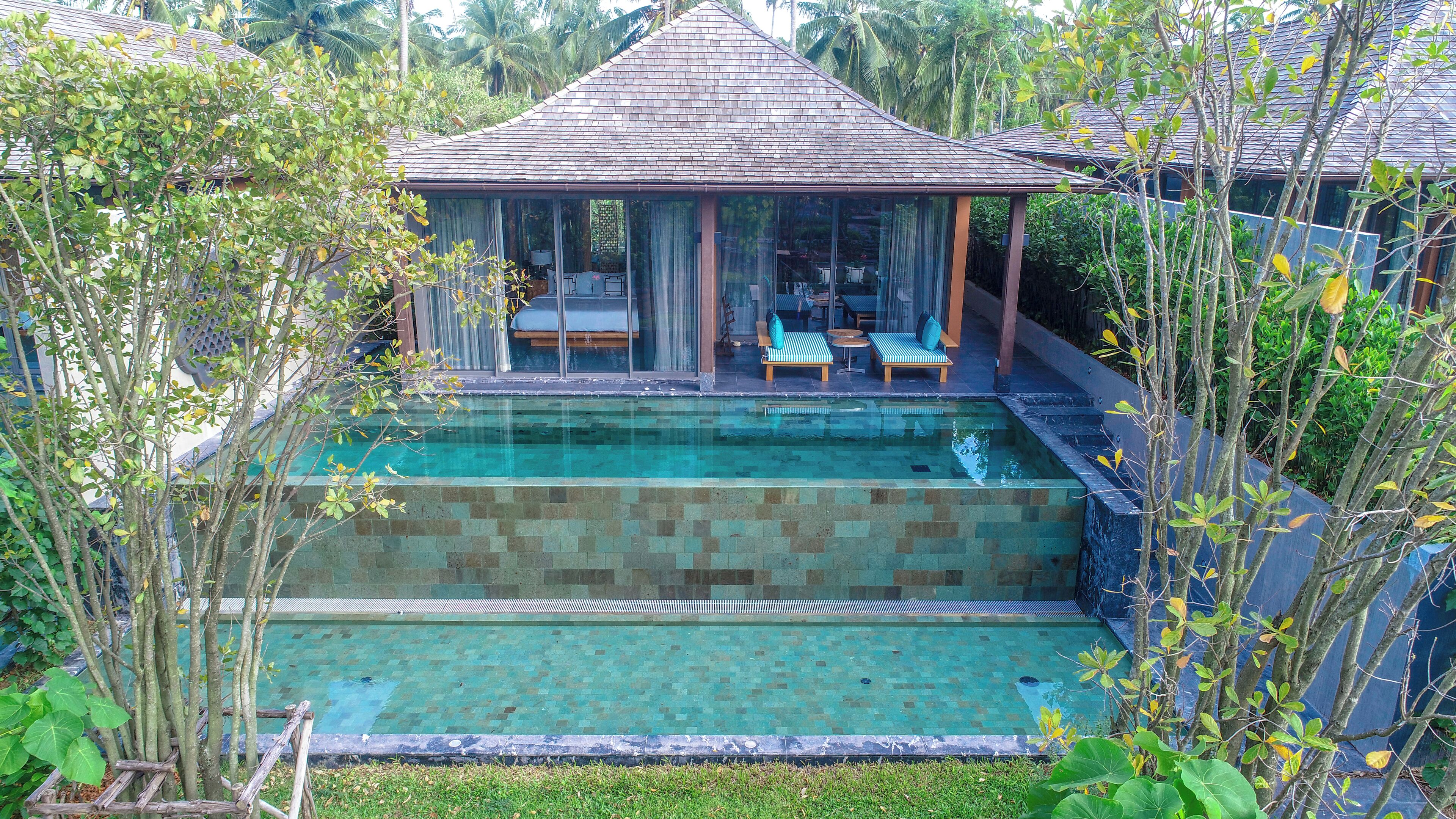 Un espace aménagé au bord de la piscine, comprenant un coin salon abrité, une piscine à eau claire et une végétation luxuriante tout autour.