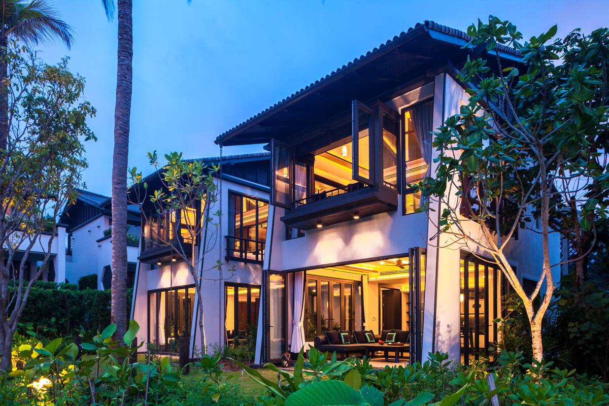 A modern two-story house with large windows and a balcony, surrounded by lush greenery.