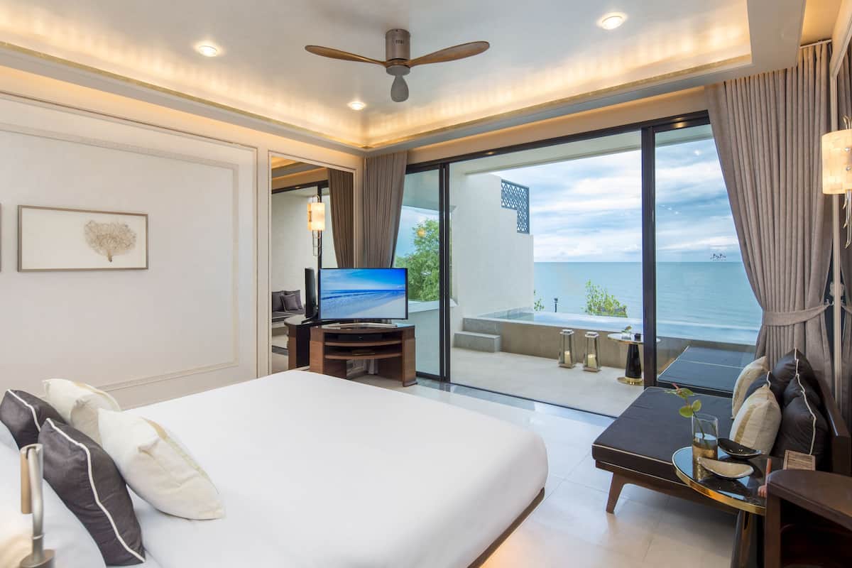 Une chambre d’hôtel avec un grand lit, un téléviseur posé sur un meuble en bois, un balcon avec vue sur la mer et un ventilateur de plafond.