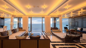 Uma sala de estar espaçosa com um sofá grande, uma mesa de centro e uma televisão. Há cortinas e vista para o oceano.