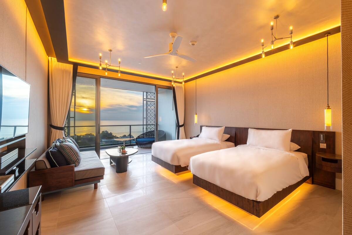 Une chambre d’hôtel moderne dotée de deux lits, d’un canapé, d’une table à manger et d’un balcon offrant une vue sur le coucher de soleil.