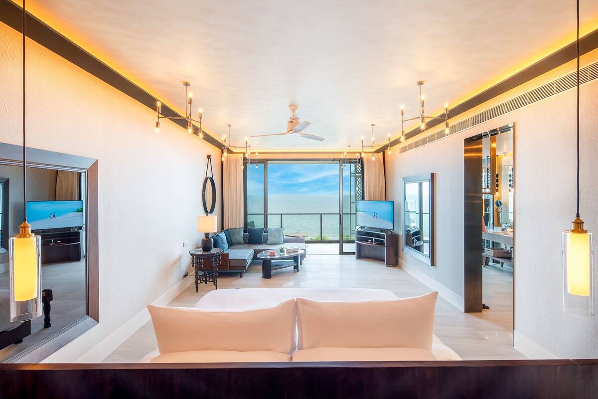 Une chambre d’hôtel moderne dotée d’un grand lit, d’un coin salon avec un canapé et une table basse, d’une télévision et d’un balcon offrant une vue sur l’eau.