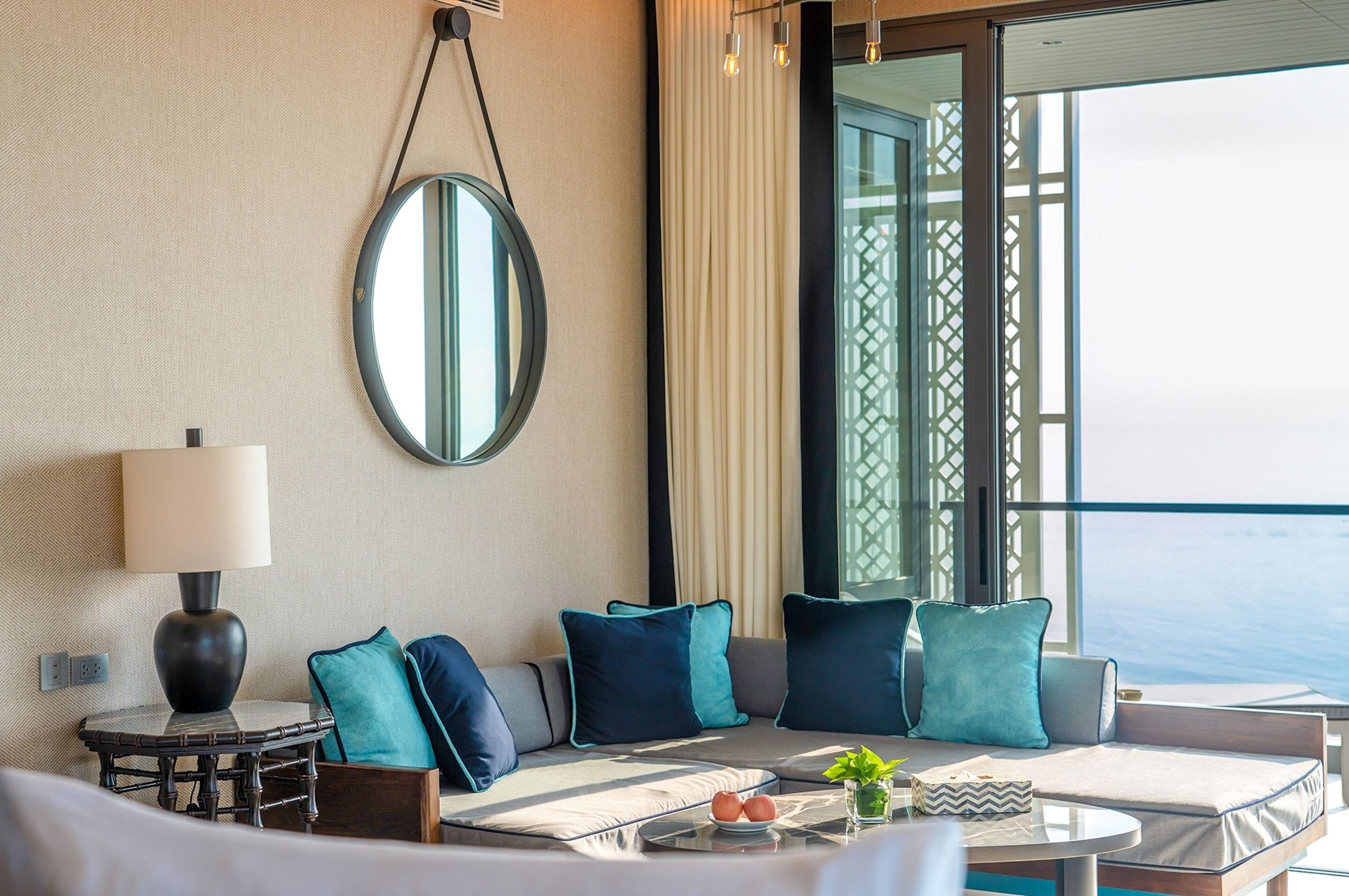 A modern hotel room with a large window, a round mirror, a sofa with teal cushions, a small table with a lamp, and a view of the sea.