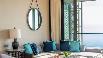 A modern hotel room with a large window, a round mirror, a sofa with teal cushions, a small table with a lamp, and a view of the sea.