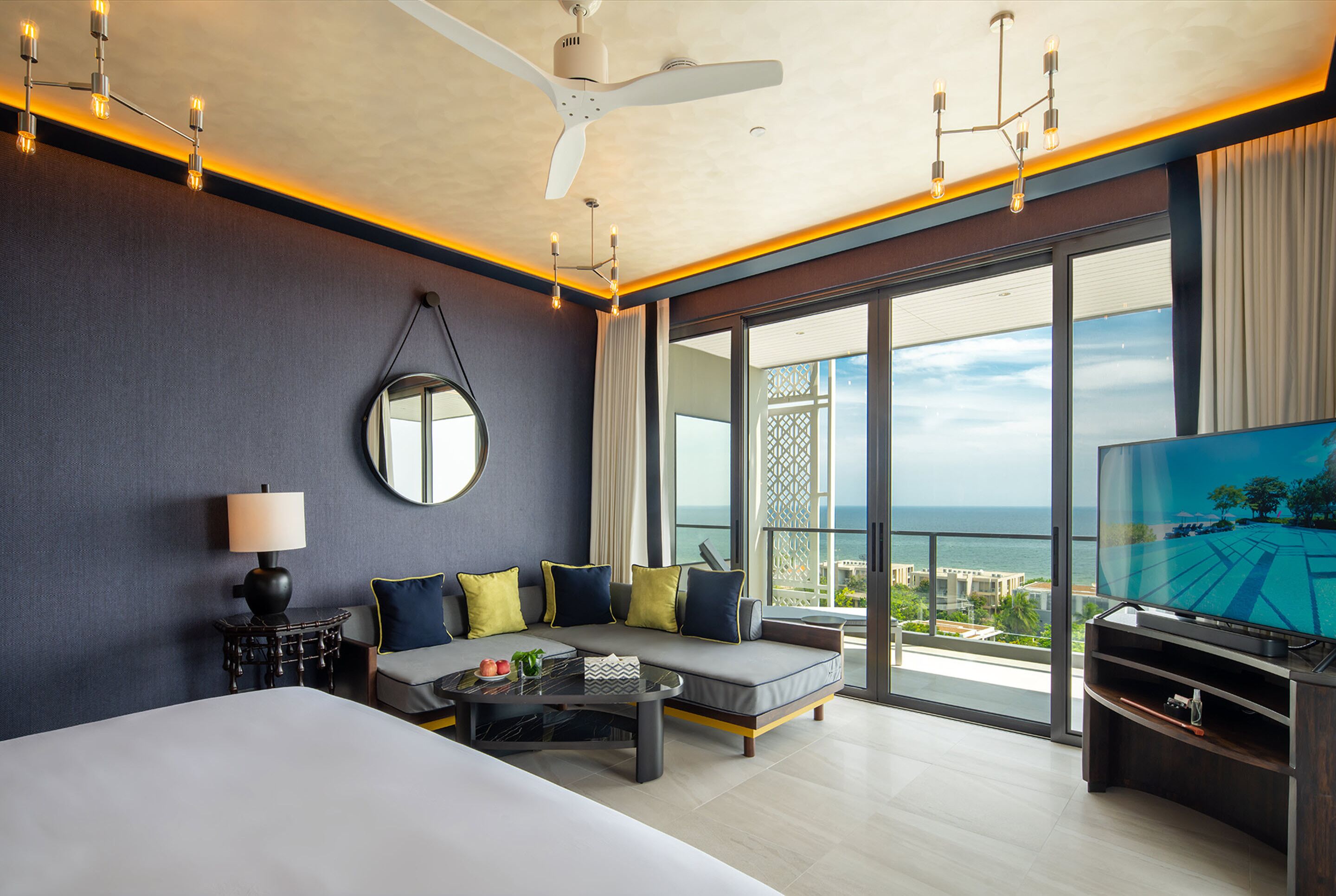 A modern hotel room with a large bed, a sofa, a TV, and a balcony with a view of the sea.