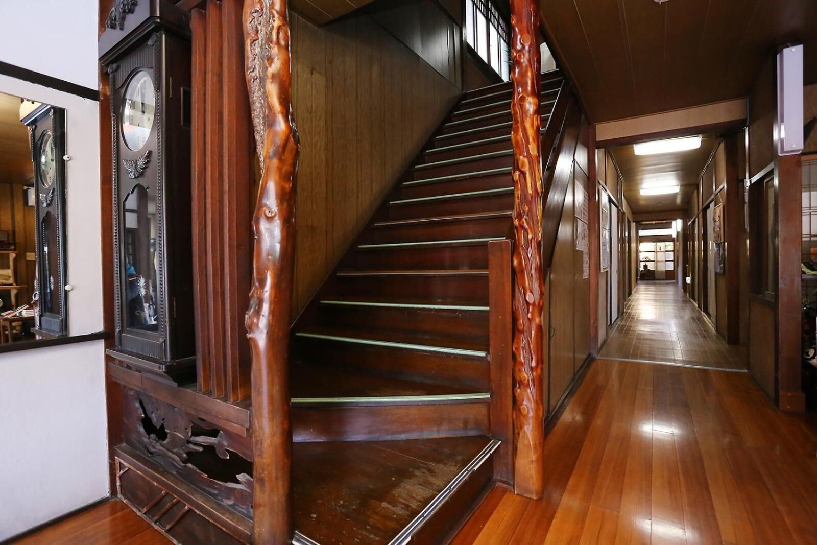 Una escalera de madera con un reloj y un espejo a la izquierda, que conduce a un pasillo con pisos de madera y puertas.
