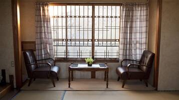 Une pièce de style japonais traditionnel, avec un sol recouvert de tatamis, deux fauteuils en cuir, une petite table ornée d’une plante et une fenêtre à motifs géométriques.