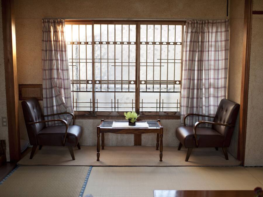 Ein traditioneller japanischer Raum mit Tatami-Bodenbelag, zwei Ledersesseln, einem kleinen Tisch mit Pflanze und einem Fenster mit Gittermuster.