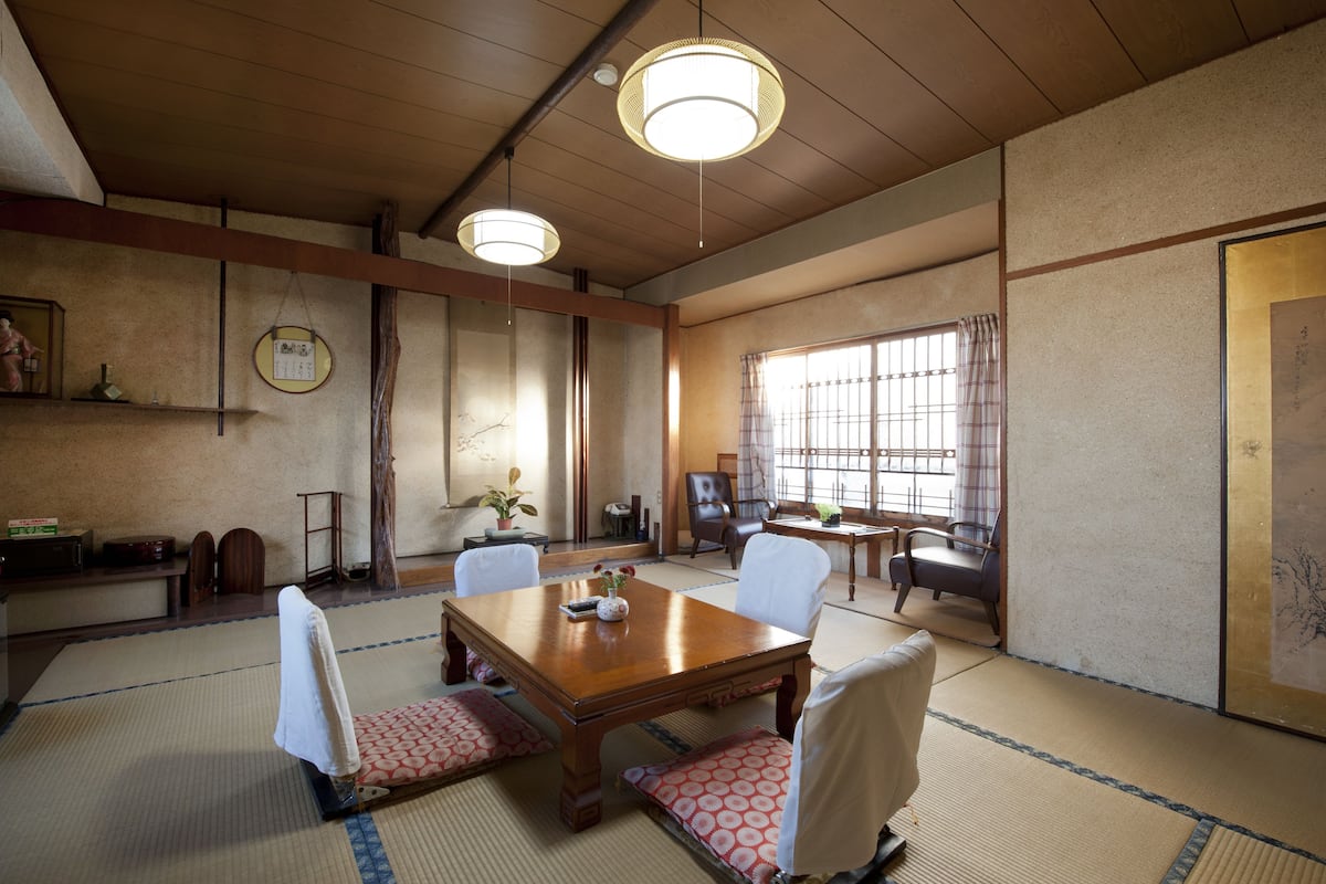 A traditional Japanese room with tatami flooring, a wooden table, and chairs. There are two hanging lamps, a window with curtains, and a sliding door.