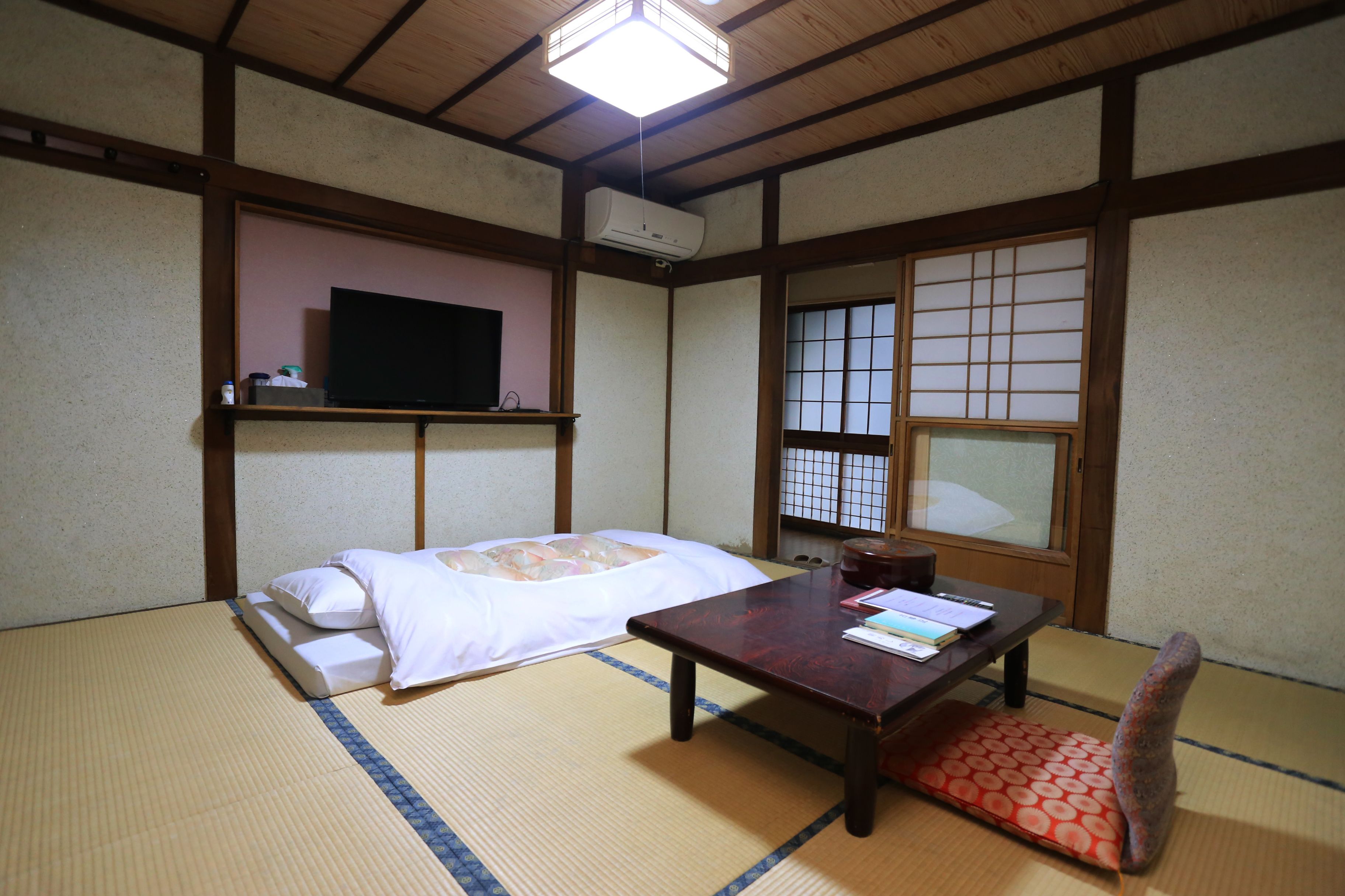 A traditional Japanese room with tatami flooring, a low table, a bed with white bedding, a television on a shelf, and a shoji screen window.