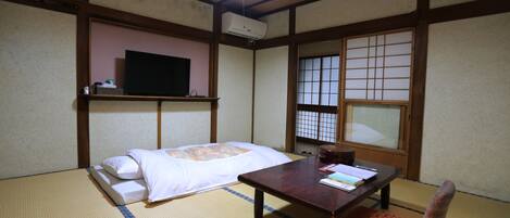 A traditional Japanese room with tatami flooring, a low table, a bed with white bedding, a television on a shelf, and a shoji screen window.