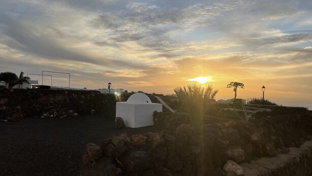 Un coucher de soleil sur la côte, avec une structure blanche au premier plan, un mur de pierre et un lampadaire.