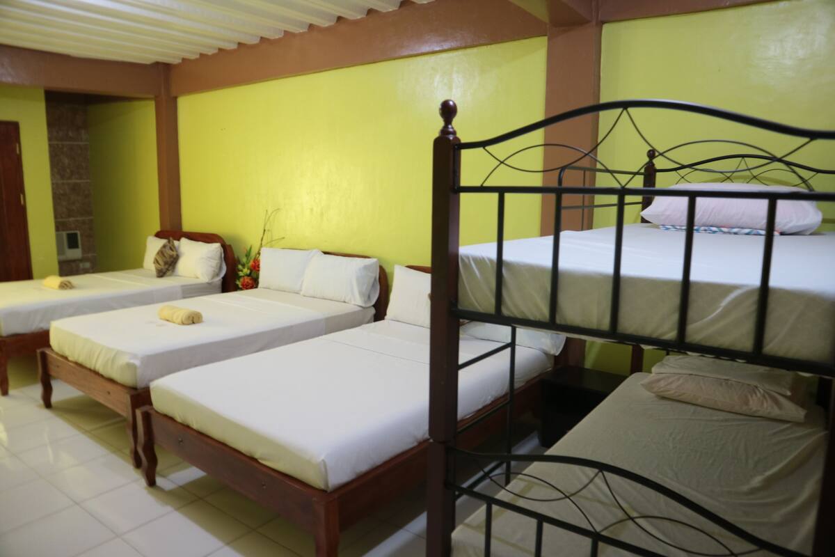 A hotel room with two single beds and a larger bed. There is a wooden headboard on the larger bed and a metal headboard on the single beds. The room has a tiled floor and a green wall.