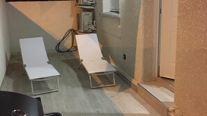 Ein kleiner Außenbereich mit einer Terrasse, zwei weißen Liegestühlen, einem Tisch und einer Glasschiebetür zu einem überdachten Eingang.
