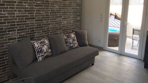 Ein Wohnzimmer mit grauem Sofa, gemusterten Kissen, einer Ziegelwand und einer Glastür, die zu einem Außenbereich mit Pool führt.