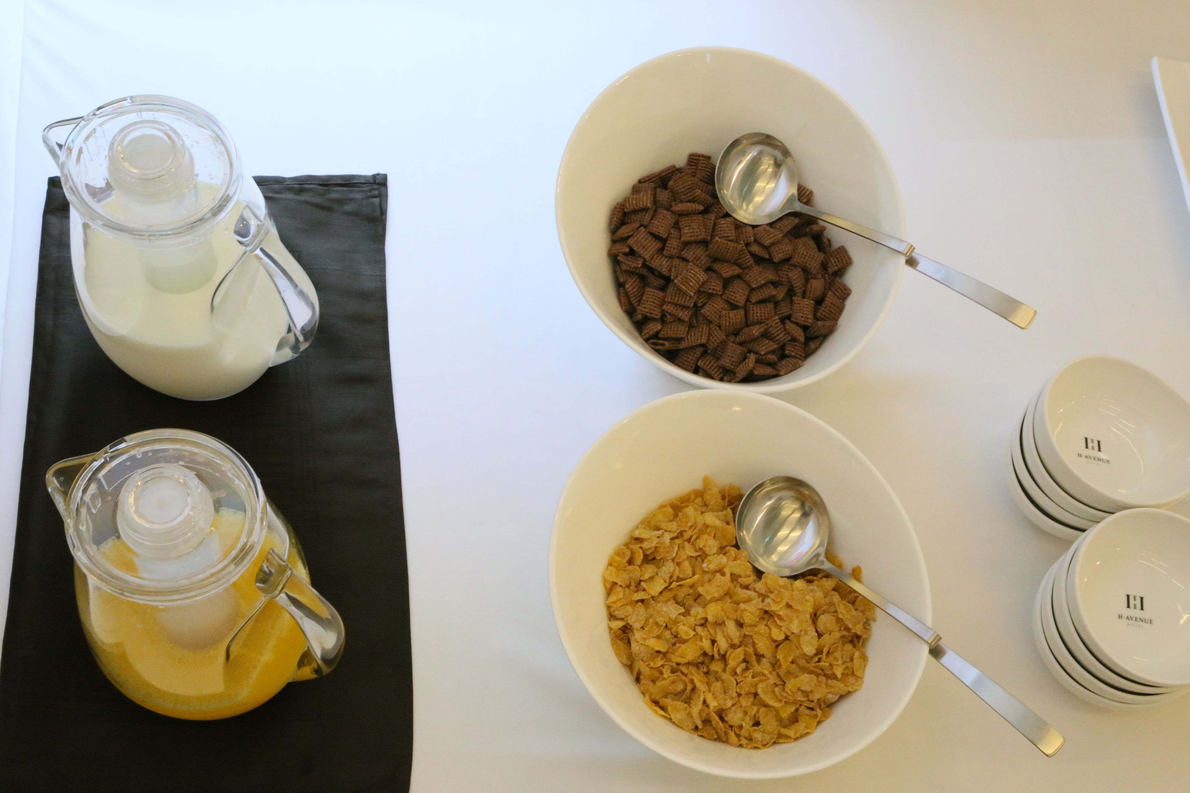 A table with three bowls containing different food items, a pitcher of milk, and a stack of bowls.