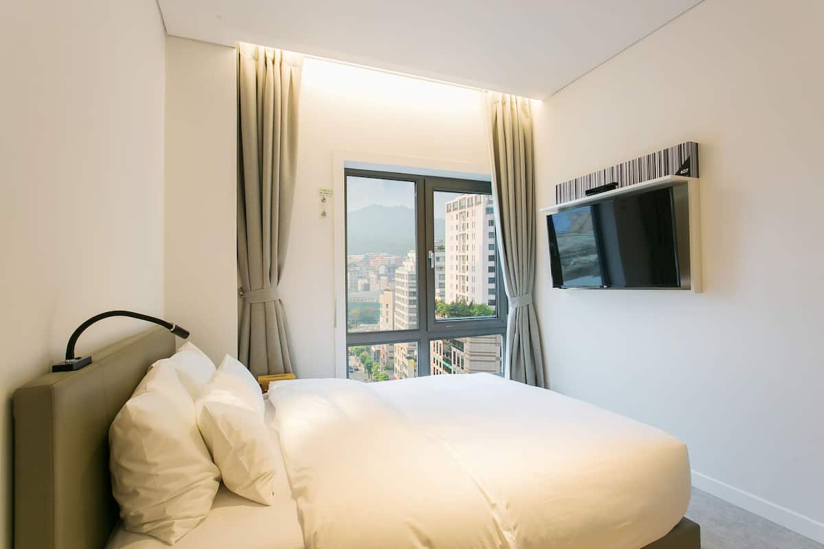 Ein Hotelzimmer mit Bett, Vorhängen, einem an der Wand montierten Fernseher und einem Fenster mit Blick auf Gebäude.