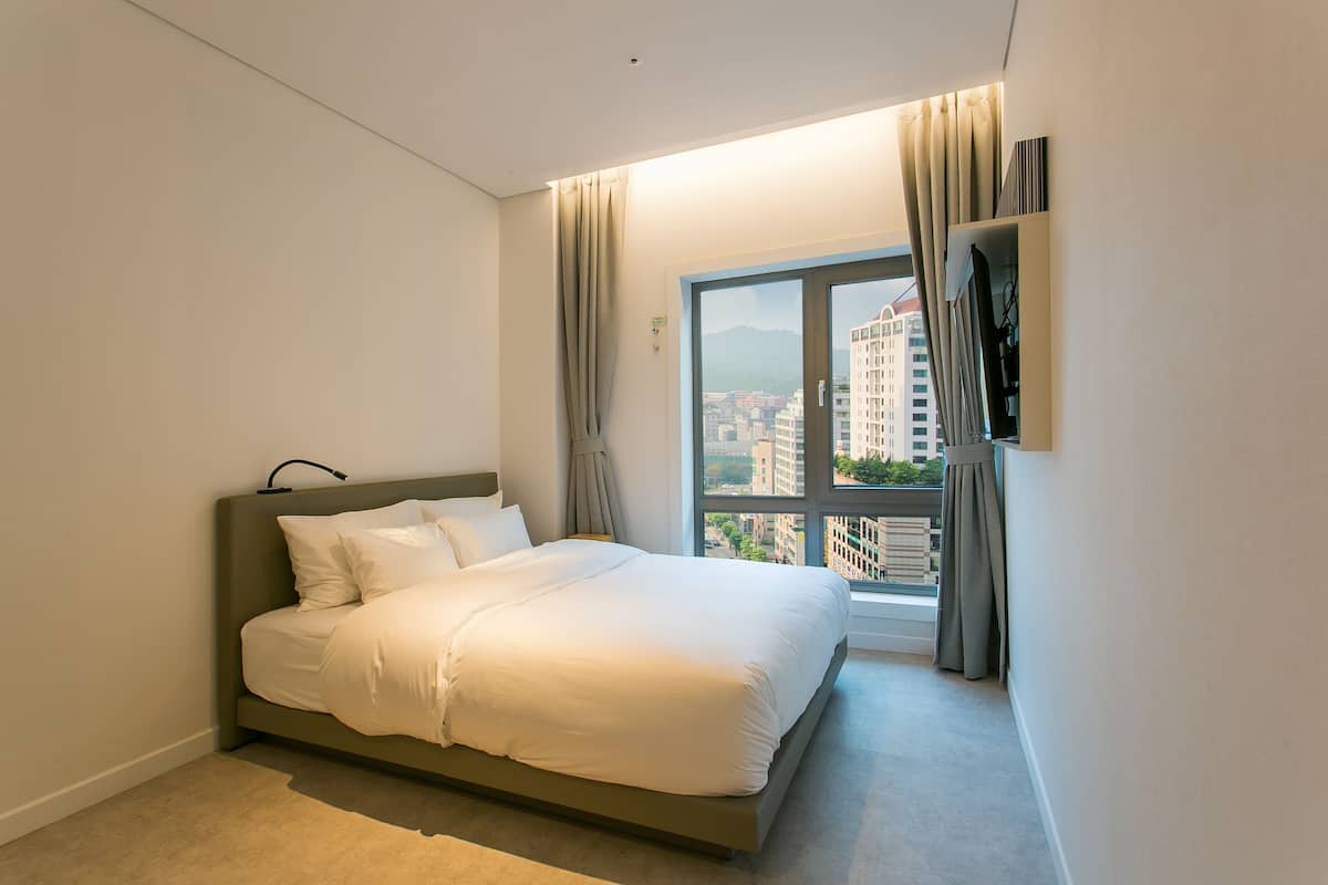Ein modernes Hotelzimmer mit einem großen Bett, einem an der Wand befestigten Fernseher und Blick auf die Stadt durch ein großes Fenster.