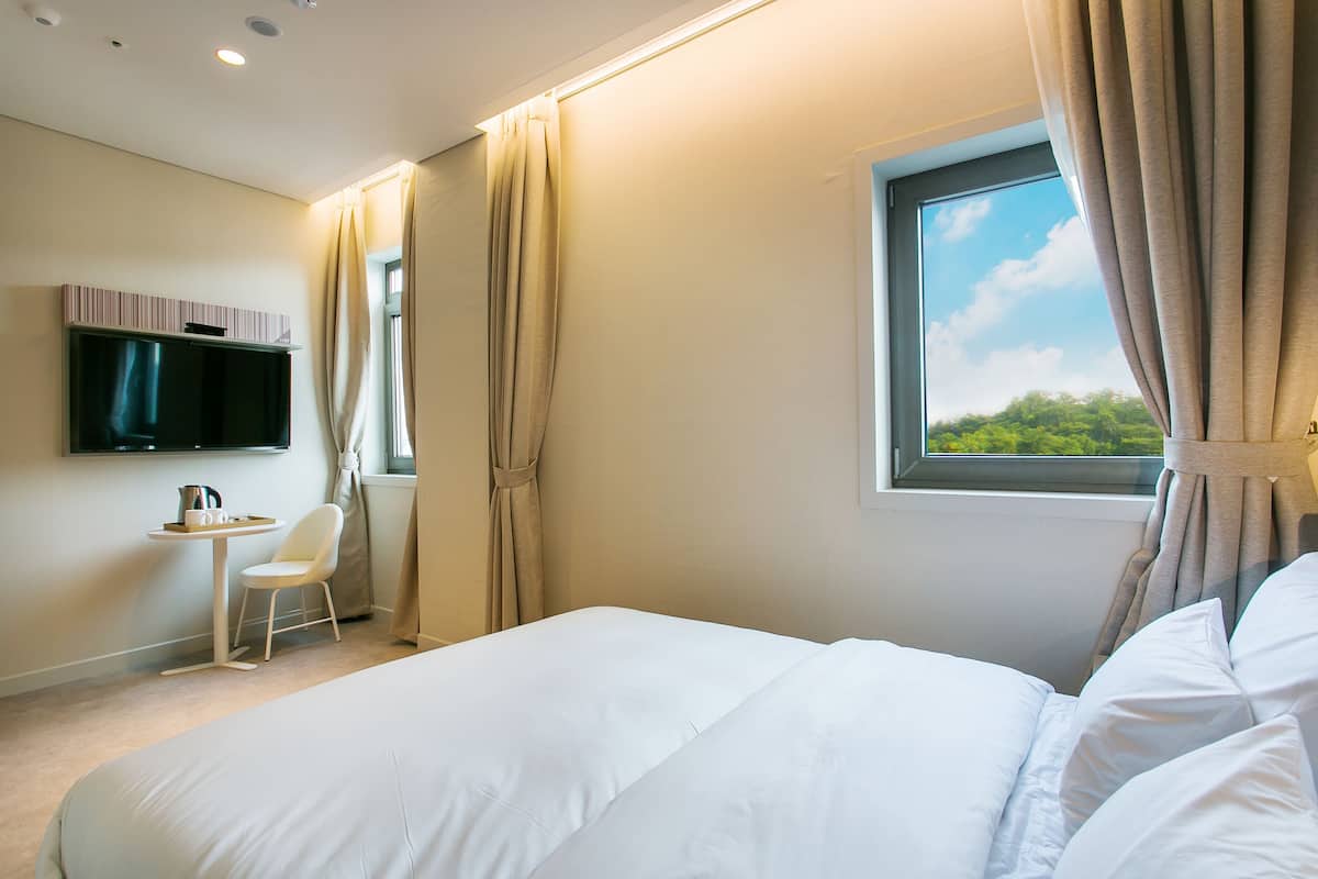 Ein Hotelzimmer mit Bett, Wandfernseher, kleinem Tisch mit Stuhl und einem Vorhangfenster.