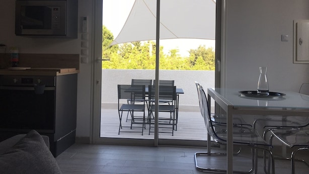 Et moderne køkken med mikrobølgeovn, et spiseområde med bord og stole, samt en skydedør til en udendørs terrasse med en hvid parasol.