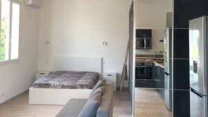 Et moderne soveværelse med seng, en grå sofa og et køkkenareal, der kan ses igennem en åben dør.