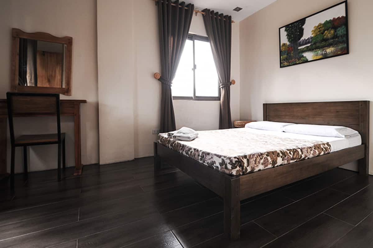 A bedroom with a wooden bed, a desk, a chair, a window with curtains, and a painting on the wall.