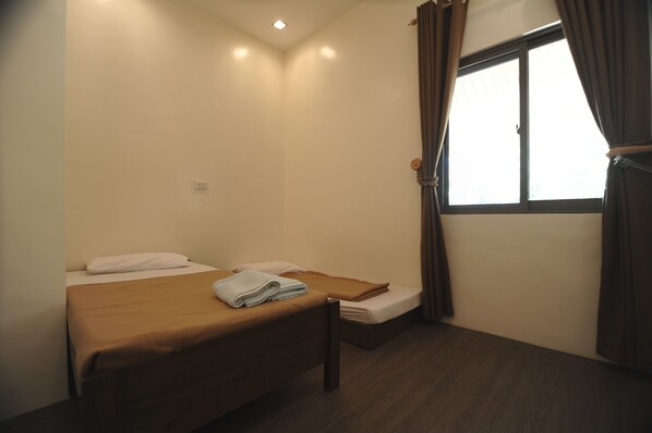 A small, single-room hotel room with a bed, a small table, and a window with curtains.