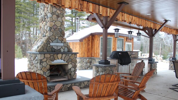 An outdoor patio with a stone fireplace, wooden chairs, and a grill.