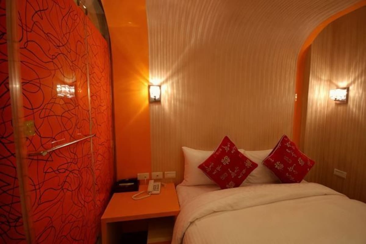 A hotel room with a curved ceiling, a bed with white bedding and red pillows, a bedside table with a phone and a lamp, and wall-mounted lights.