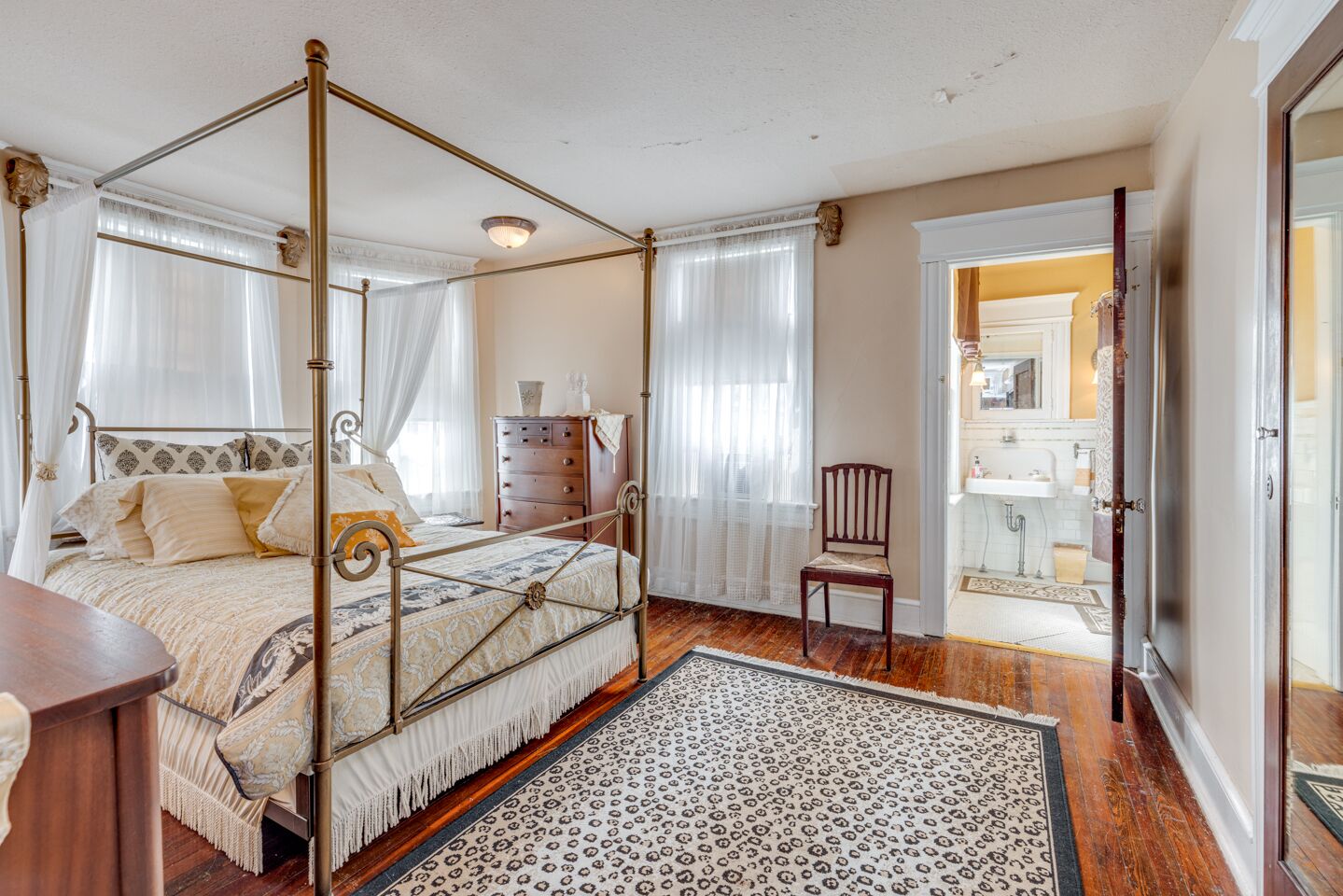 A four-poster bed with a canopy, a wooden dresser, a chair, and a bathroom visible through an open door.