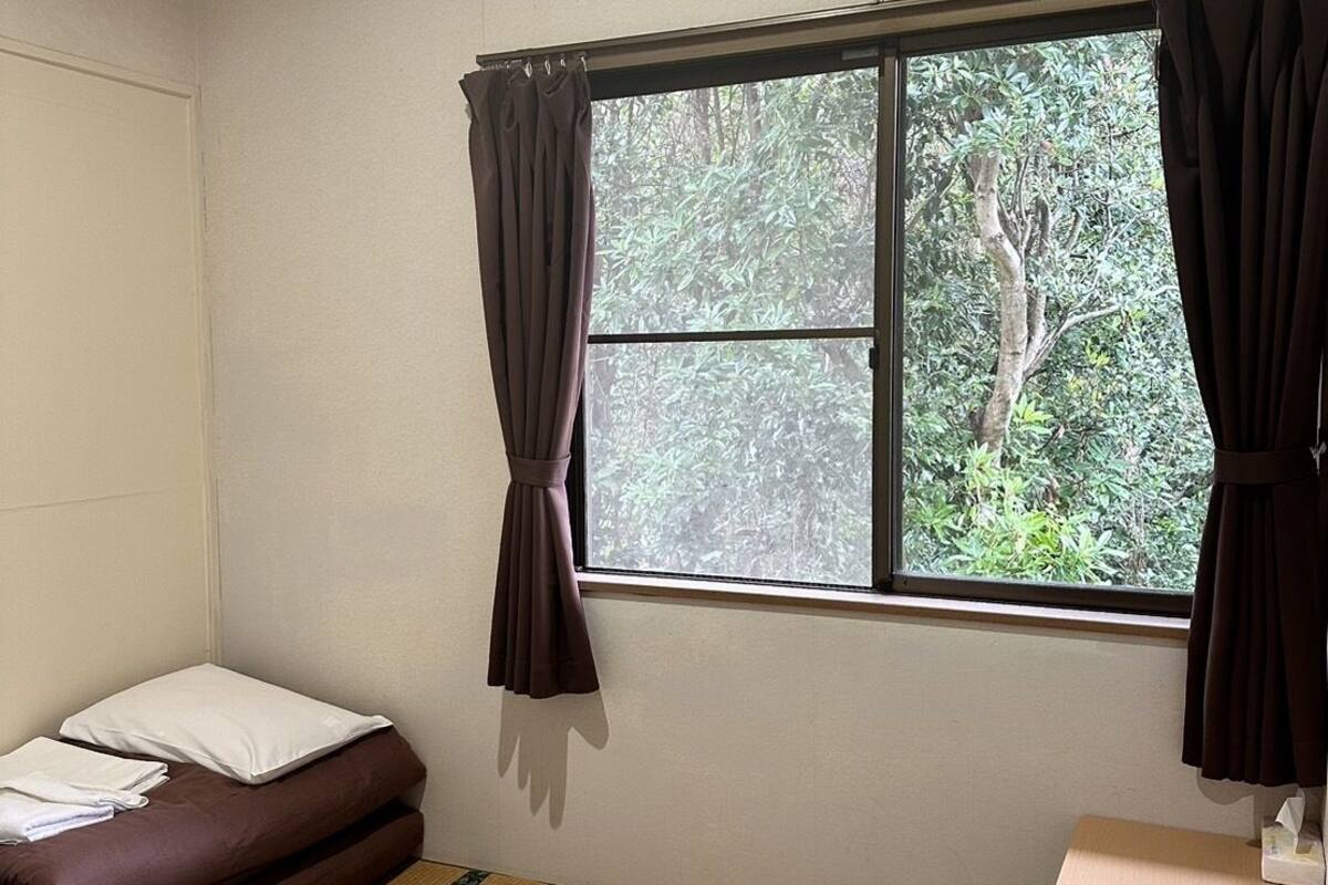A small room with a bed, a window with curtains, an air conditioner, and a small table with a trash bin.