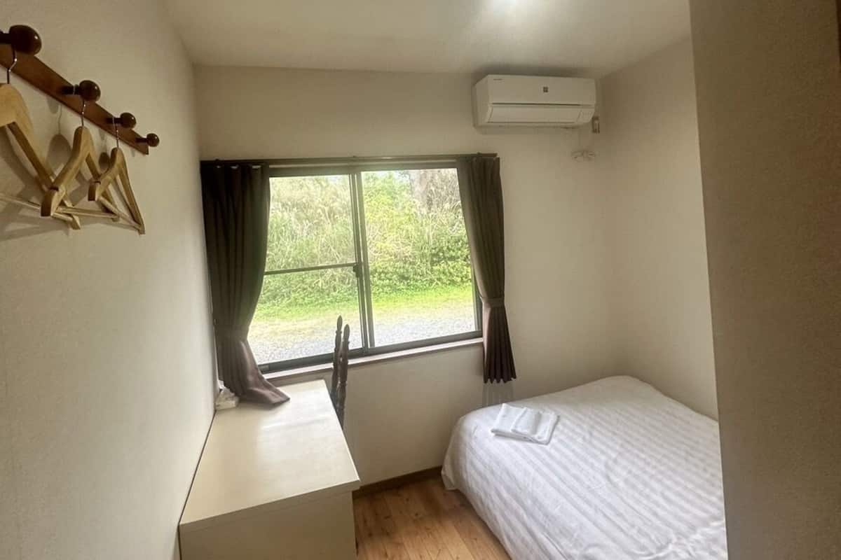 A small, single-room accommodation with a wooden floor, a bed, a desk, and a window offering a view of greenery outside.