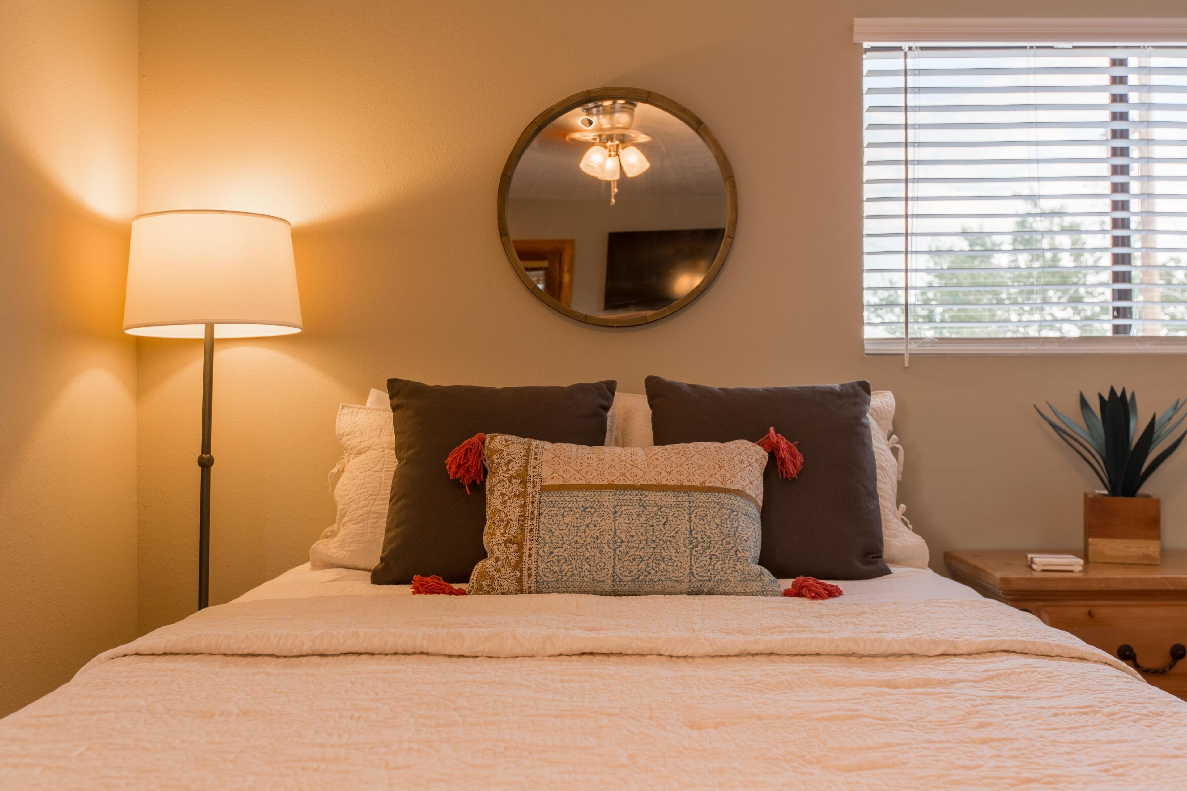 Una cama bien hecha con edredón blanco y almohadas decorativas, una lámpara de noche, un espejo redondo en la pared y una ventana con persianas.