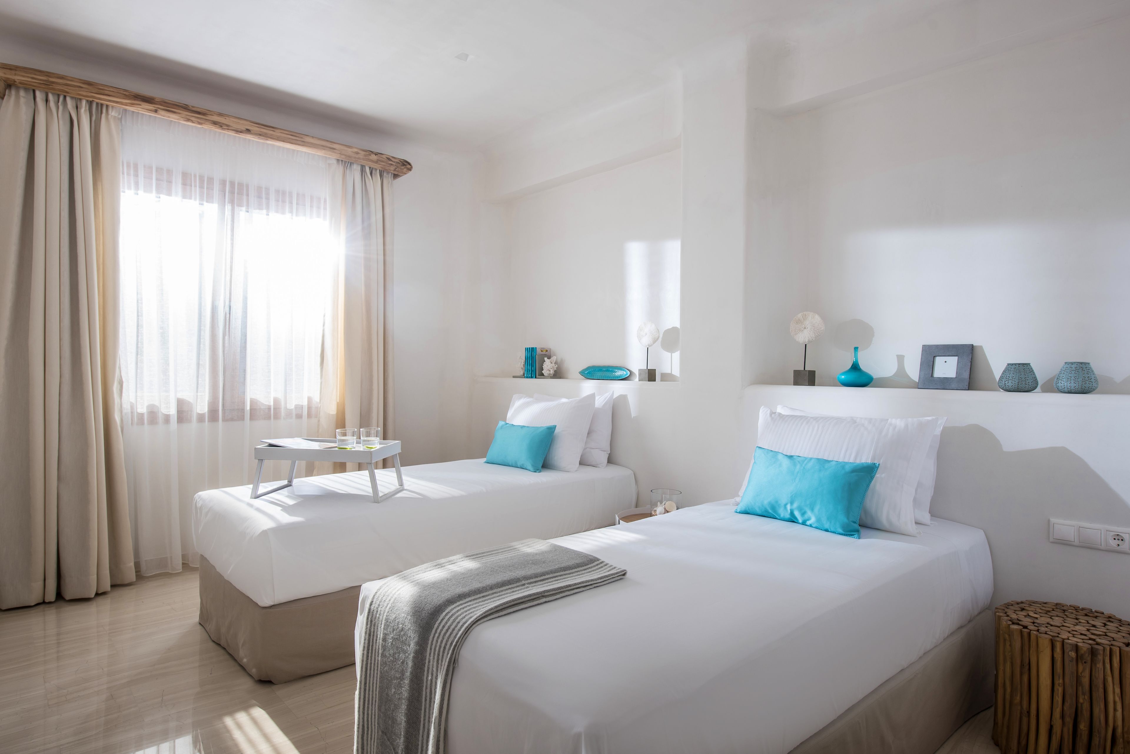 A hotel room with two beds, a small table, a mirror, and a shelf with decorative items.