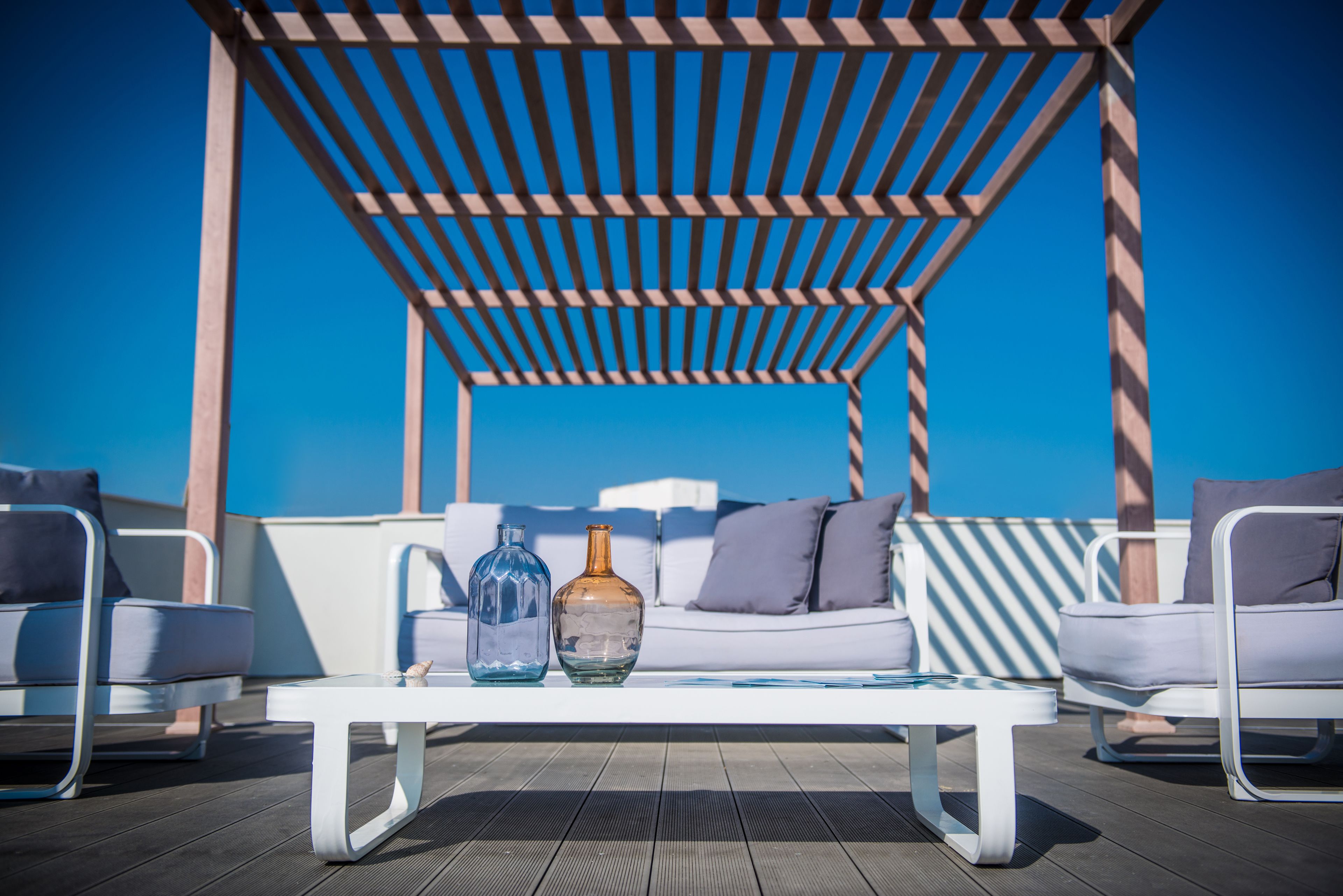 A sunlit outdoor seating area with a white table, two glass bottles, and a white sofa with cushions.