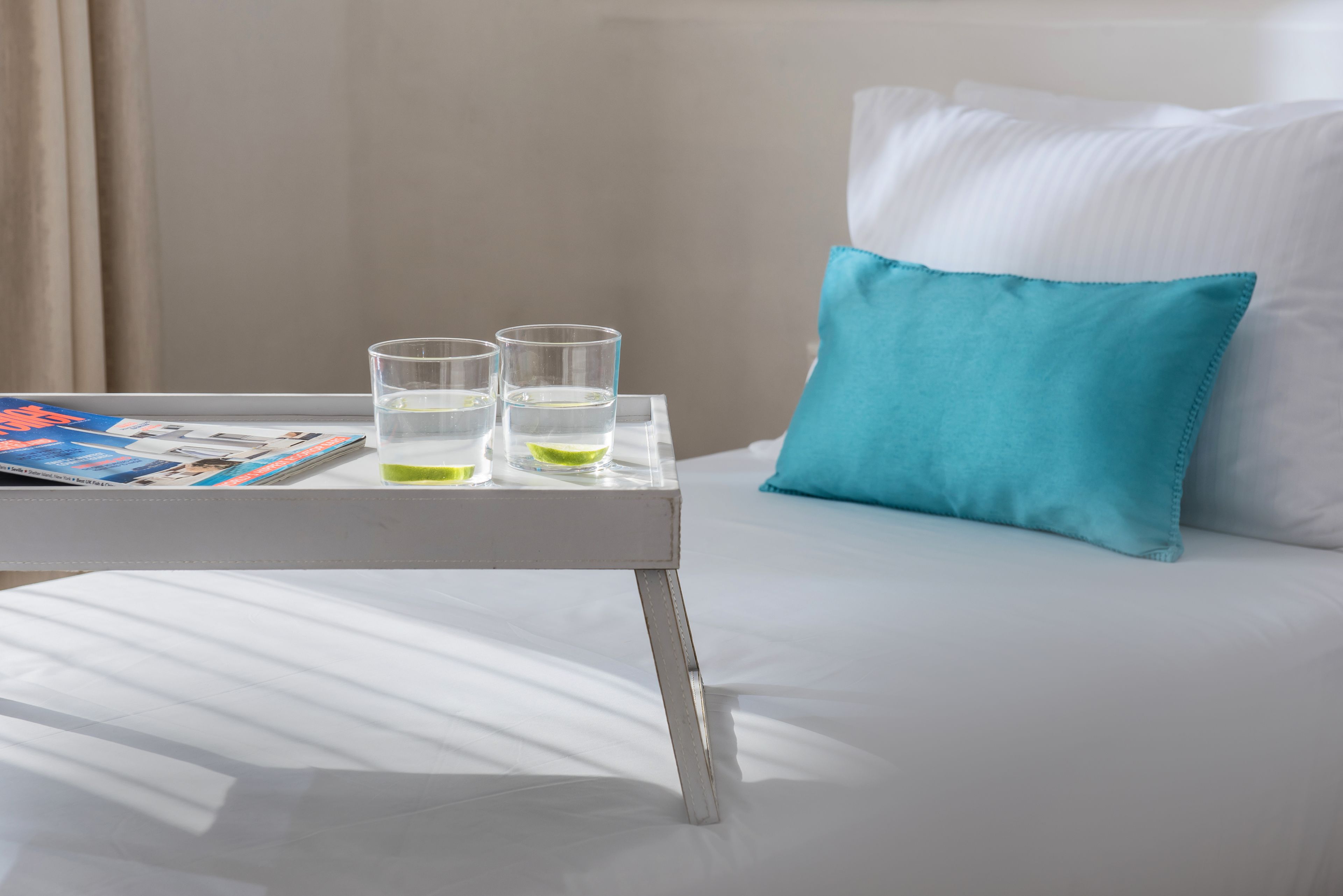 A neatly made bed with white sheets and a blue pillow, a small table with two glasses of water and a magazine, and a window with curtains.