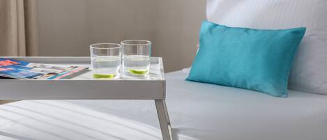 A neatly made bed with white sheets and a blue pillow, a small table with two glasses of water and a magazine, and a window with curtains.