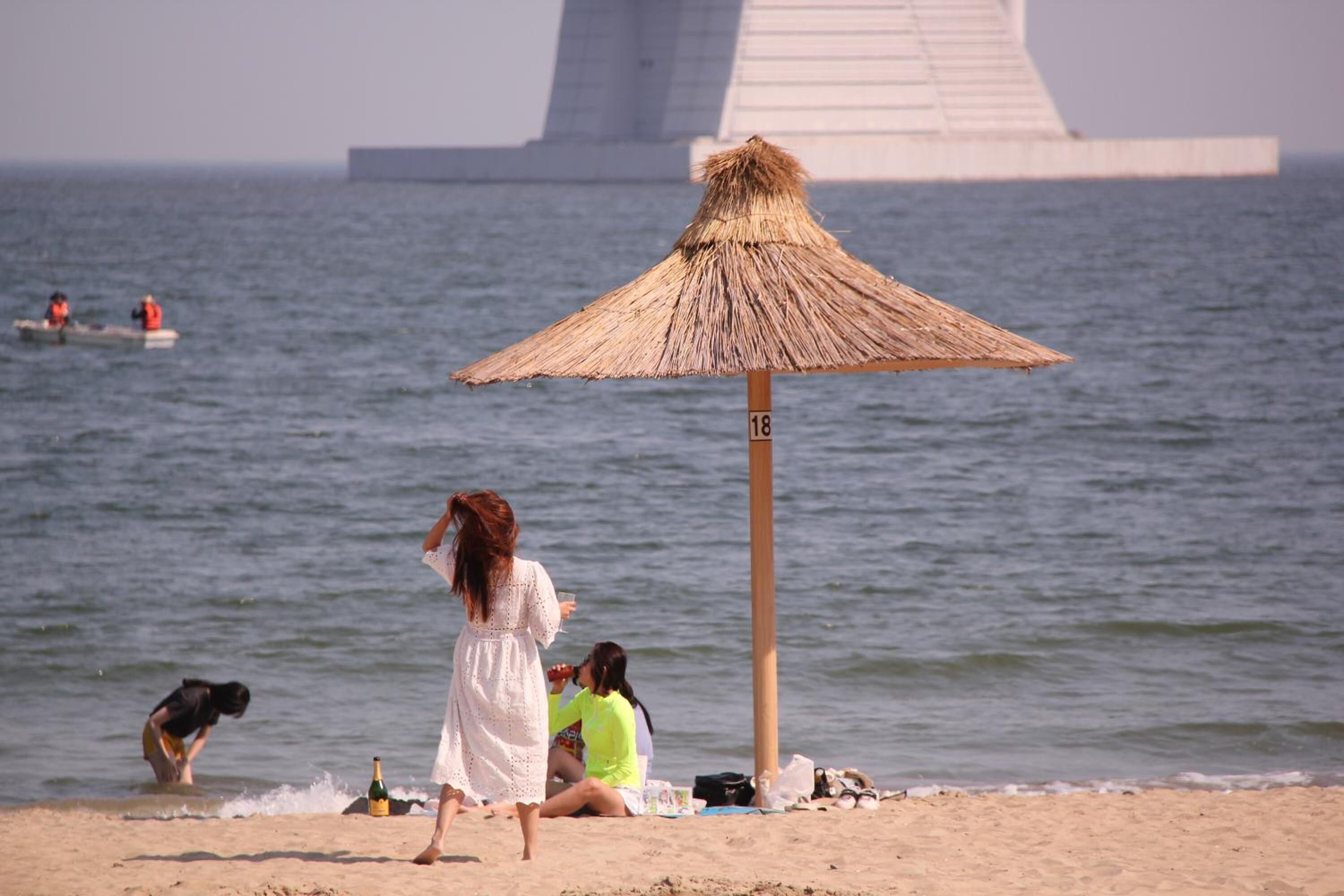 A beach with a straw umbrella, people relaxing, and a large structure in the background.