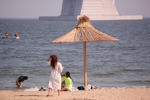 A beach with a straw umbrella, people relaxing, and a large structure in the background. - Wood House Hotel (Busan)
