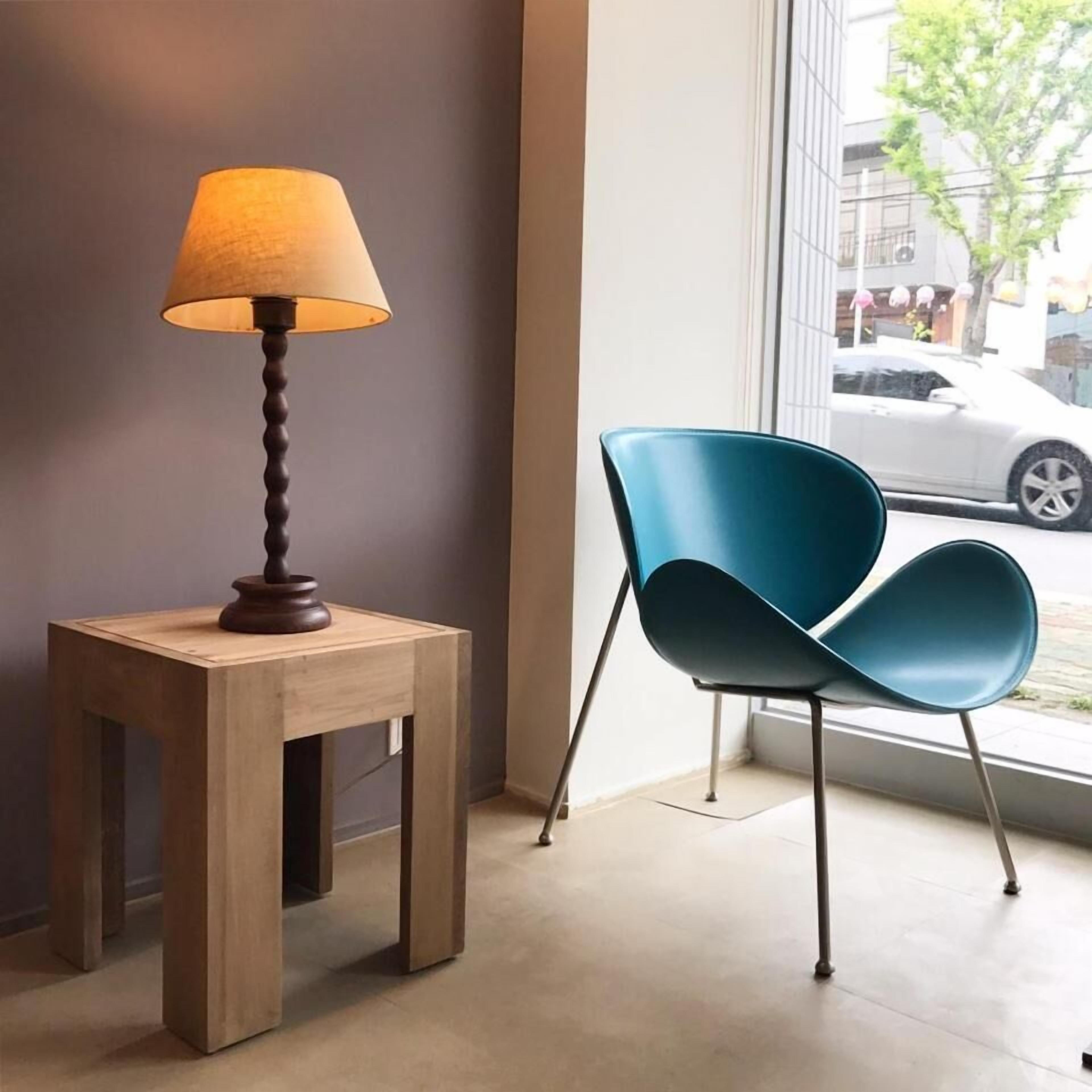 A modern interior with a wooden side table holding a lamp, a blue chair, and a large window offering a view of the street.
