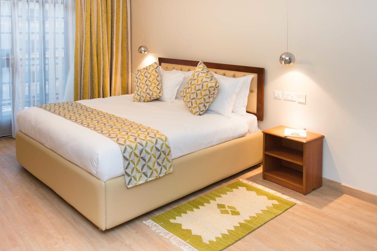 Una cama bien tendida con almohadas estampadas, una mesita de noche con lámpara y una alfombra sobre piso de madera.