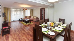 Un comedor con una mesa de madera dispuesta para cuatro personas, rodeada de sillas. Un sofá y una mesa baja se observan al fondo.